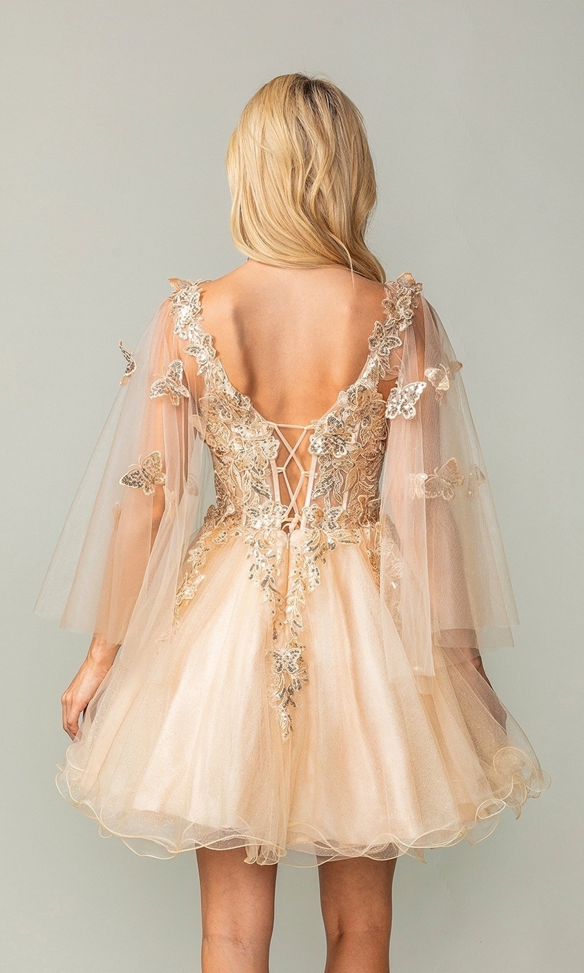 A woman with long blonde hair models the Butterfly-Cape Short A-Line Homecoming Dress 3393 in peach tulle, featuring sequin appliques, sheer butterfly-embellished sleeves, and an open lace-up back.